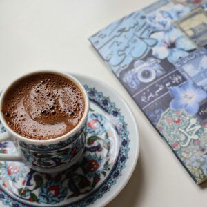 Close-up of Turkish coffee in ornate cup with Arabic journal on the side.